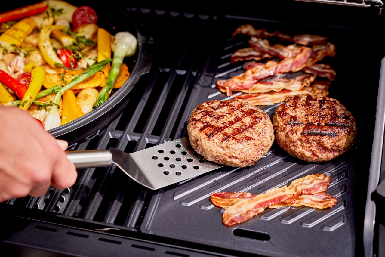 Grillplatte mit gegrillten Burger-Patties und Speck, daneben eine Pfanne mit Gemüse, aus der Perspektive von oben.