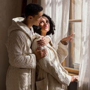 Zwei Personen in cremefarbenen Kapuzenmänteln stehen nebeneinander und schauen aus einem Fenster. Die Mäntel haben ein gestricktes Muster und wirken gemütlich. Die Szene ist von der Seite aufgenommen.