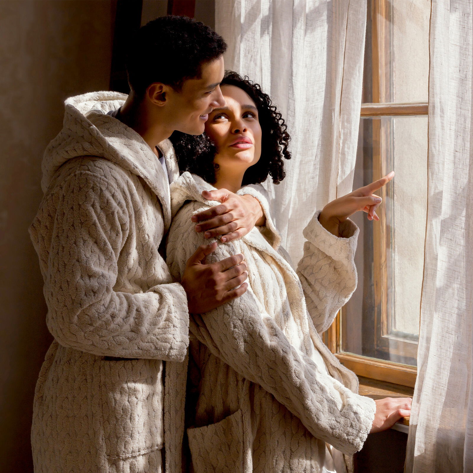 Zwei Personen in cremefarbenen Kapuzenmänteln stehen nebeneinander und schauen aus einem Fenster. Die Mäntel haben ein gestricktes Muster und wirken gemütlich. Die Szene ist von der Seite aufgenommen.