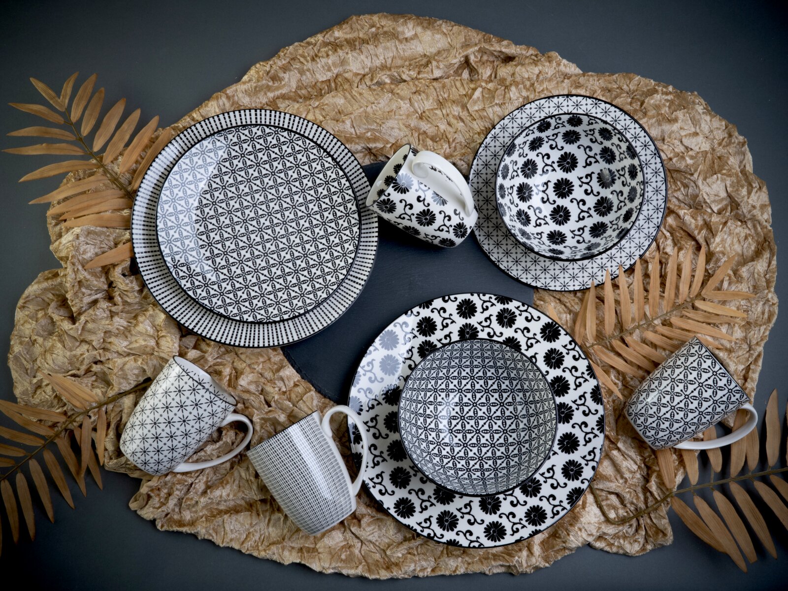 Schwarz-weiß gemustertes Geschirr-Set mit Tellern, Schalen und Tassen auf dekorativem Papier und Trockenblättern, Draufsicht (Flatlay)