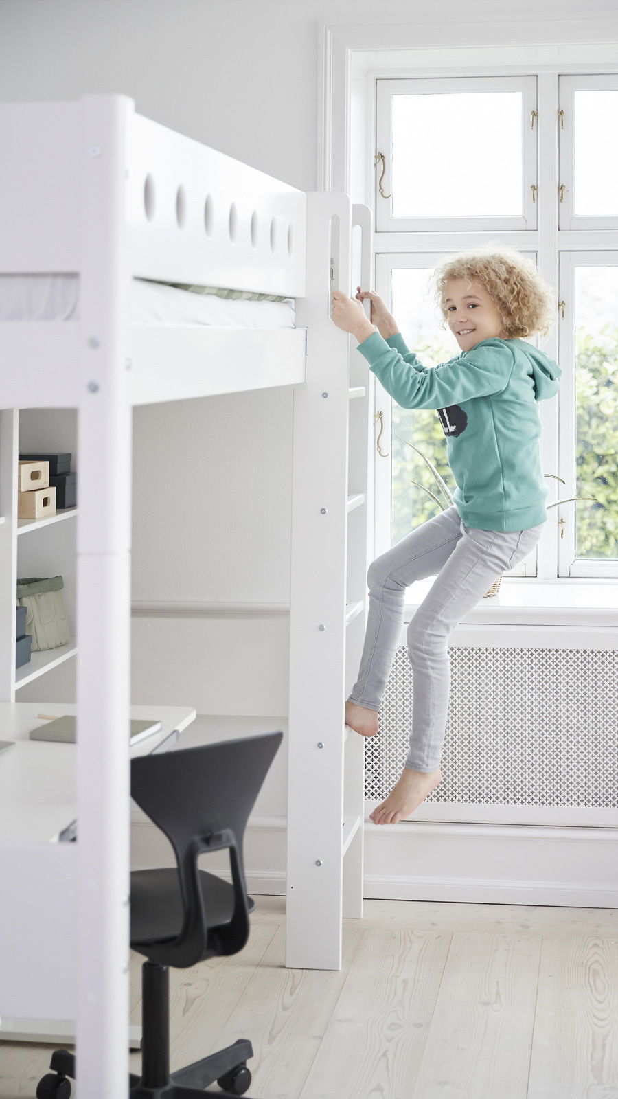 Ein Kind klettert eine weiße, senkrechte Leiter eines Hochbetts hinauf. Im Hintergrund ist ein Schreibtisch mit einem schwarzen Stuhl zu sehen. Die Perspektive zeigt das Hochbett von der Seite.