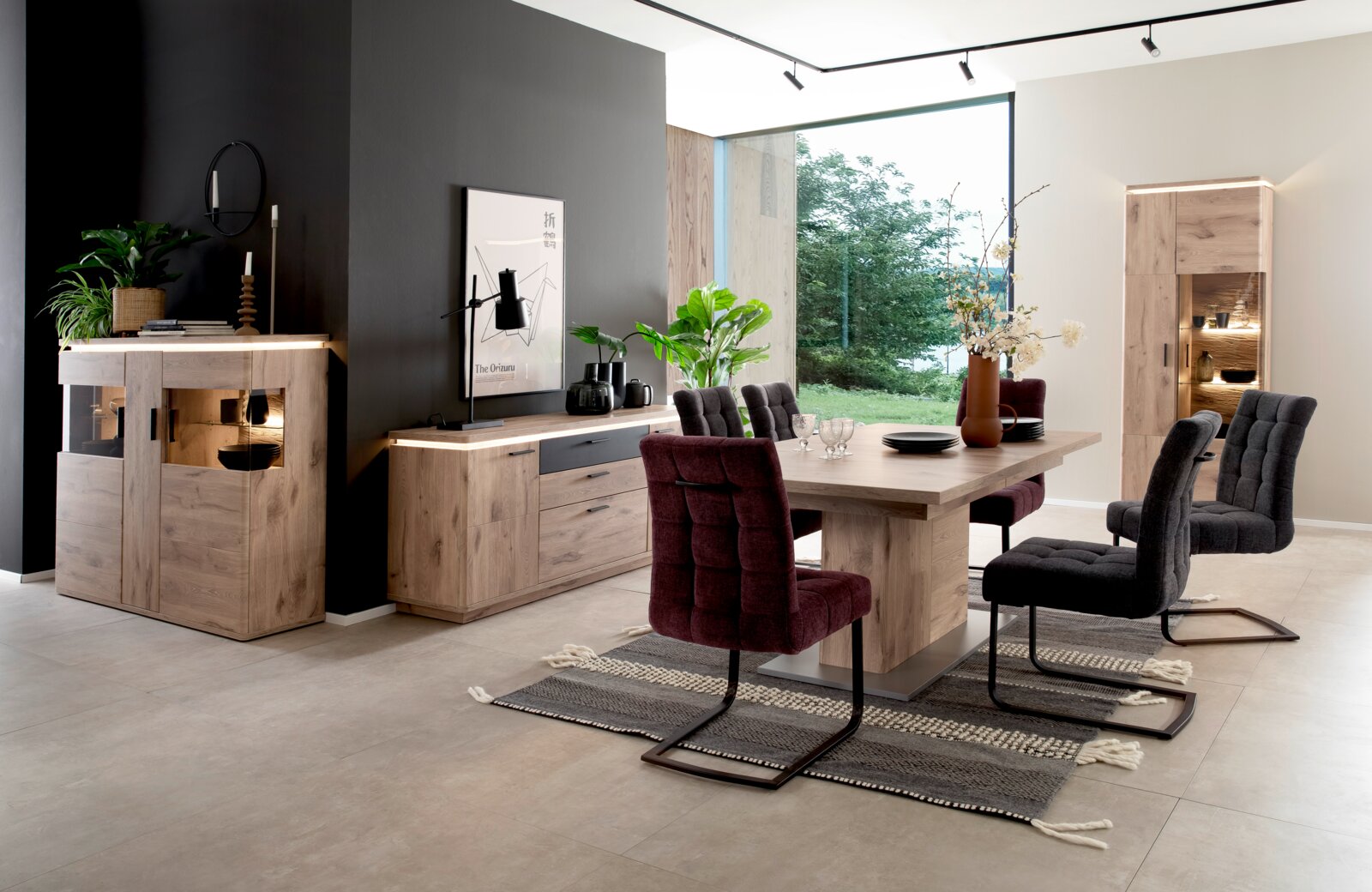 Moderne eetkamerset in houtlook met een grote eettafel, gestoffeerde schommelstoelen in bordeauxrood en antraciet, en bijpassende dressoirs en een vitrinekast met verlichting; perspectief: licht verhoogd, frontaal groothoekbeeld vanaf links.