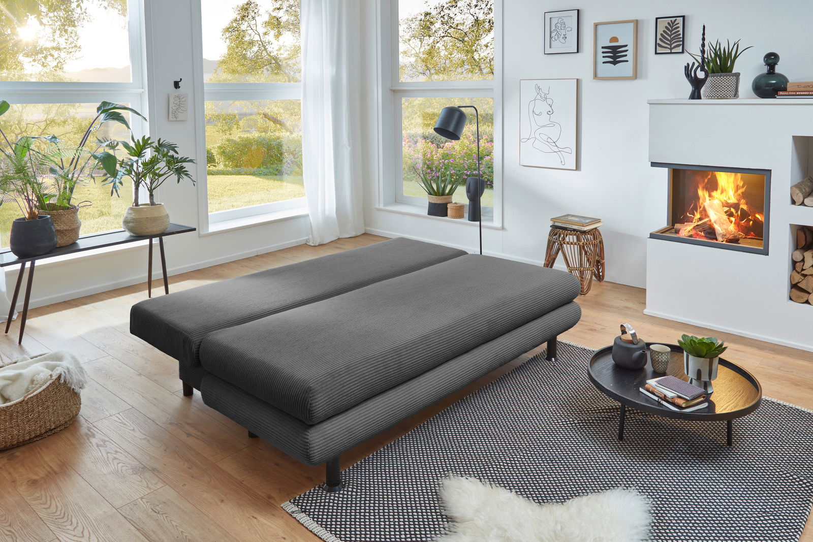 Graues Funktionssofa in einem modernen Wohnzimmer, von der Seite betrachtet, mit großen Fenstern und einem Kamin im Hintergrund.