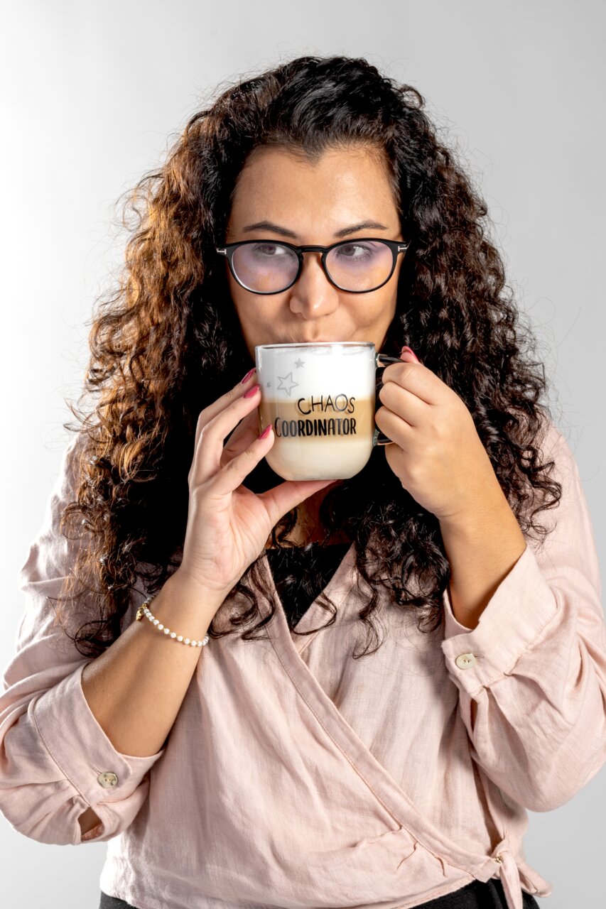 Frau mit lockigem Haar und Brille hält eine Tasse mit der Aufschrift 'CHAOS COORDINATOR' in beiden Händen, frontal betrachtet.