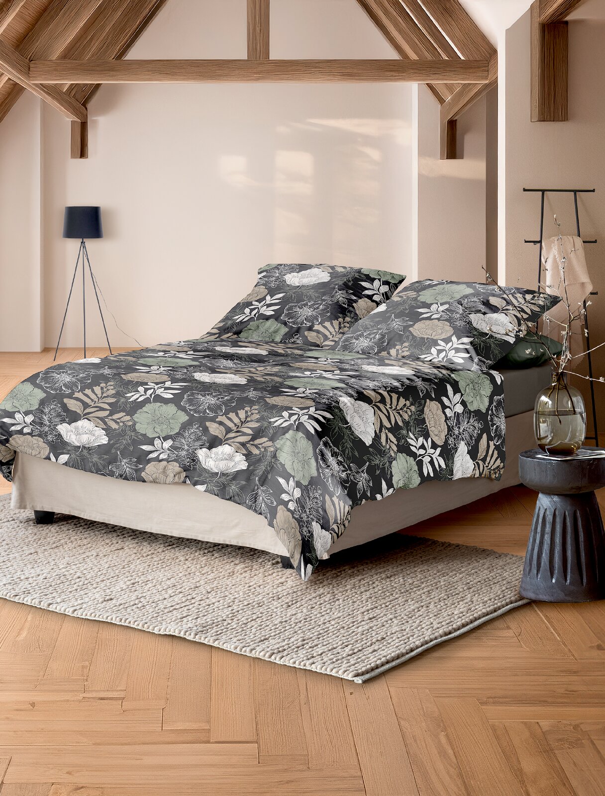 Schuin vooraanzicht van een stijlvol bed met donkergrijs dekbedovertrek en bijpassende kussenslopen, versierd met grote bloemen en bladeren in wit, beige en zachtgroen.