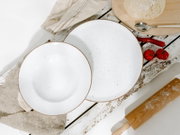 Draufsicht auf ein weißes, gesprenkeltes Keramik-Teller- und Schüssel-Set mit braunem Rand auf rustikalem Holztisch, neben Teig, Löffel, Chili und Tomaten