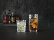 Twee kristallen glazen in etnisch design, het ene met een heldere drank en een schijfje limoen, het andere met een donkere drank, op een donker oppervlak tegen een wazige achtergrond met flessen. Zijaanzicht.