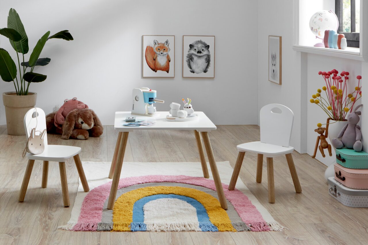 Heller Kinderzimmer-Spielbereich mit weißem Kindertisch und zwei passenden Stühlen mit Holzbeinen auf Regenbogen-Teppich, frontal aus leicht erhöhter Perspektive aufgenommen.