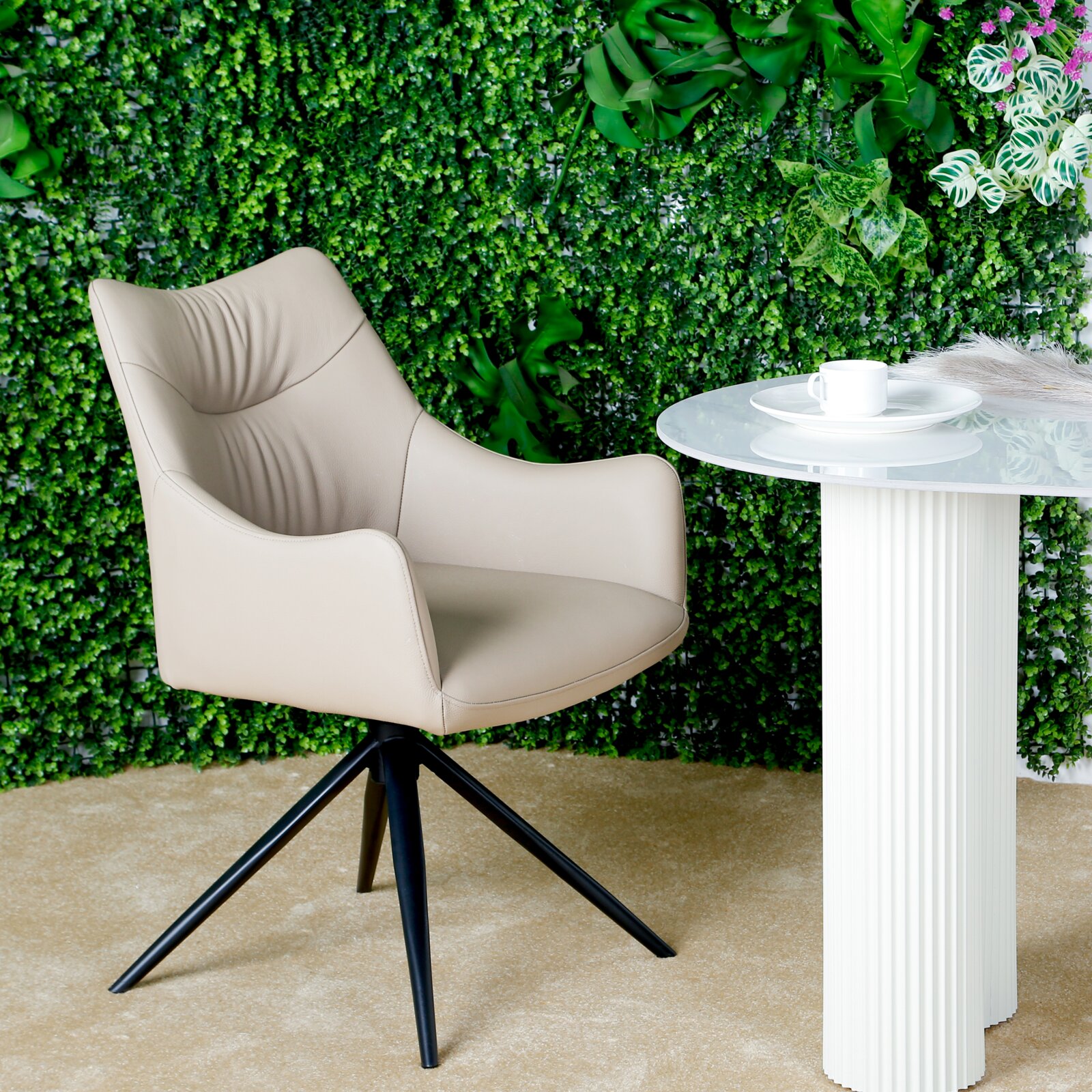 Novel Fauteuil BELFORT Beige fauteuil met zwart metalen frame, voor een groene plantenwand en naast een witte tafel, gezien vanuit de zijkant.