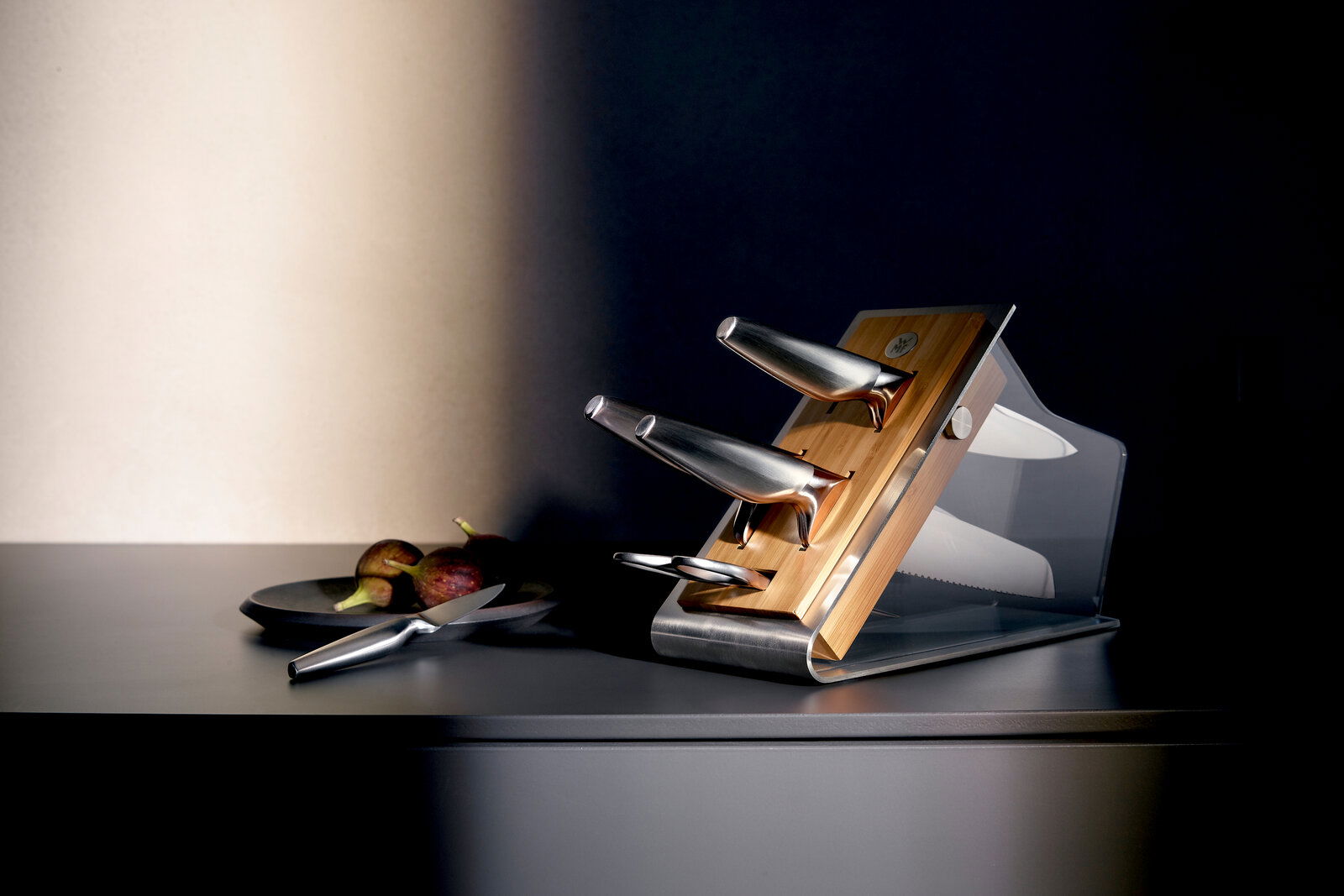 Moderner Messerblock aus Holz und Metall mit drei Messern, seitliche Perspektive, auf einer dunklen Küchenarbeitsplatte.