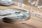 Stapel von drei Tellern aus dem 18-teiligen Portofino-Set, seitliche Perspektive, mit Besteck und Serviette auf einem Holztisch arrangiert.