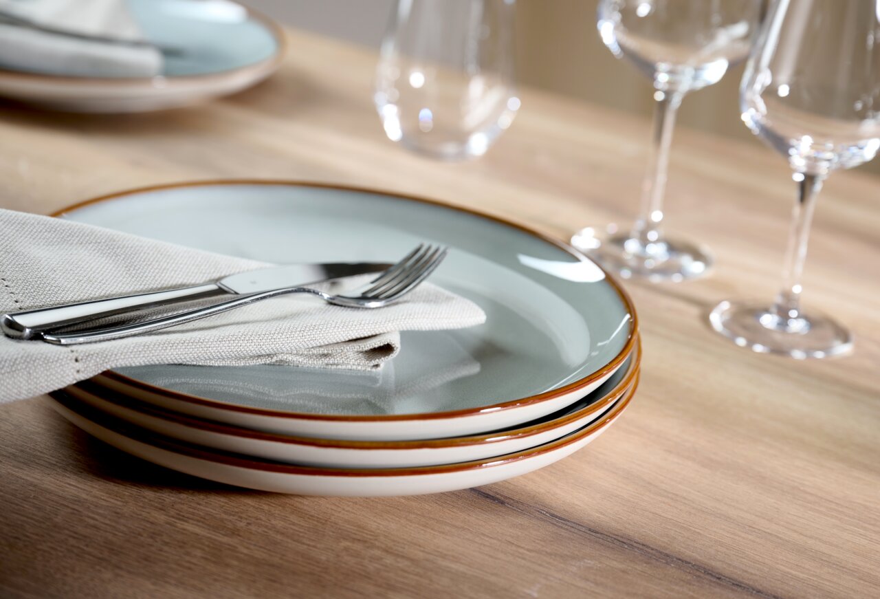 Ritzenhoff & Breker Servies 18-delig PORTOFINO Stapel von drei Tellern aus dem 18-teiligen Portofino-Set, seitliche Perspektive, mit Besteck und Serviette auf einem Holztisch arrangiert.