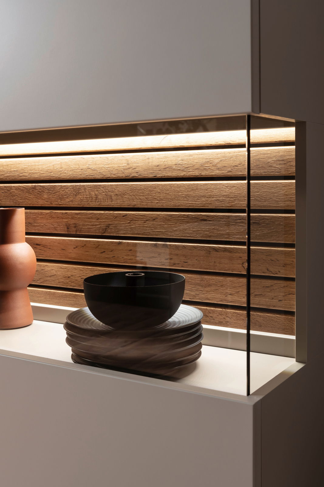 Nahaufnahme eines Highboards mit Holzrückwand und integrierter Beleuchtung, dekoriert mit einer schwarzen Schale auf gestapelten Tellern und einer terrakottafarbenen Vase, seitliche Perspektive.
