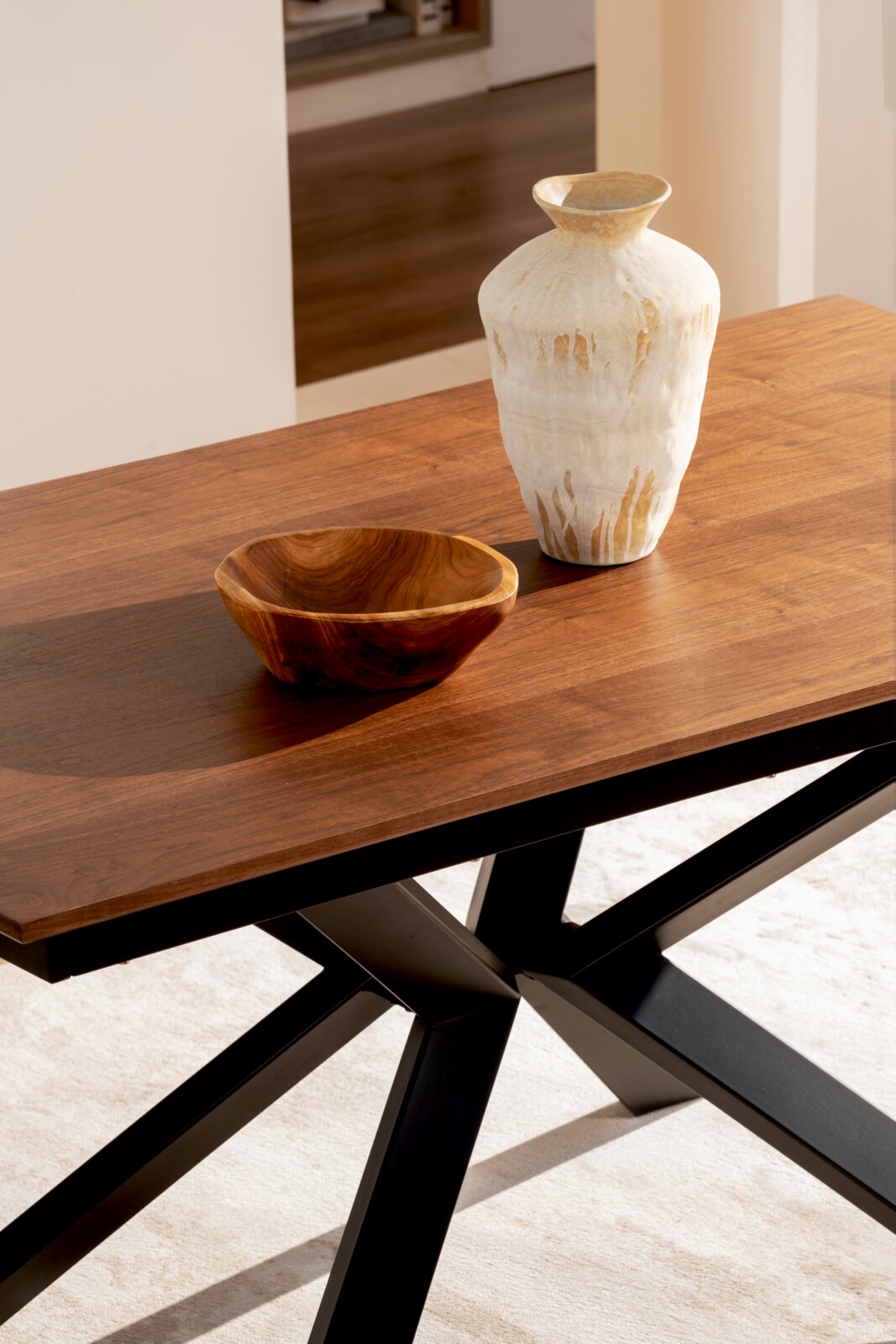 Schuin van boven gefotografeerde moderne houten eettafel met warmbruine tafelblad en zwart metalen kruisframe, gestyled met een witte keramische vaas en een houten schaal