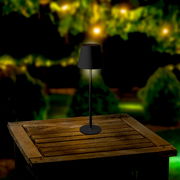 Solar Tischleuchte auf einem Holztisch im Freien, bei Nacht mit unscharfem, beleuchtetem Hintergrund, seitliche Perspektive