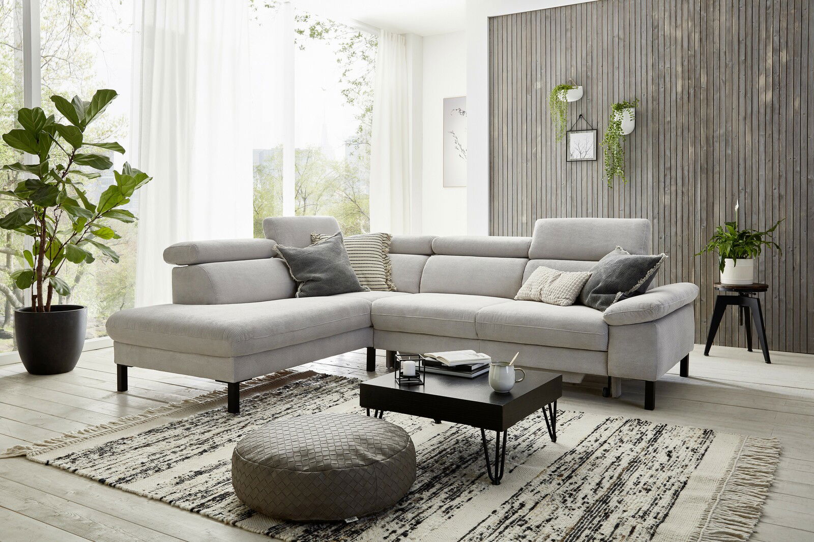Moderne Eckgarnitur in hellem Wohnzimmer mit großen Fenstern, Pflanzen und Holzwand, seitliche Perspektive