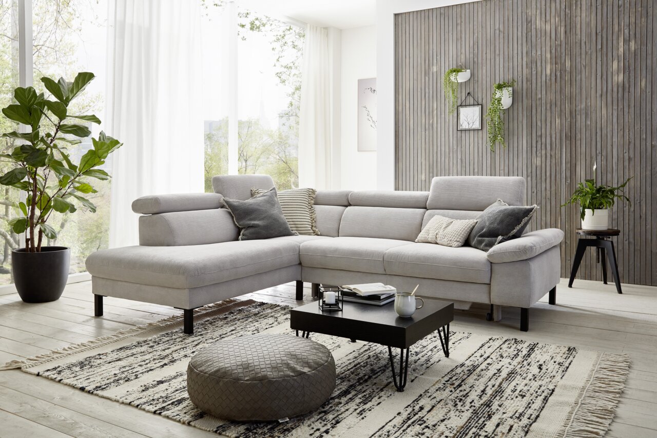 Moderne Eckgarnitur in hellem Wohnzimmer mit großen Fenstern, Pflanzen und Holzwand, seitliche Perspektive