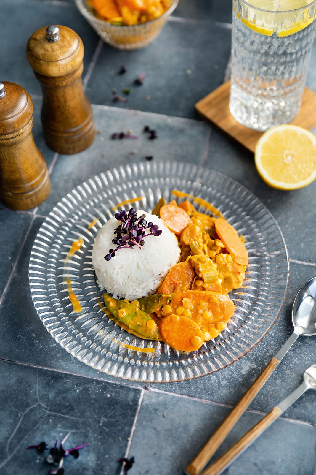 Glasierter Teller aus der Vogelperspektive mit Reis und Gemüse-Curry, umgeben von Pfeffermühlen, einem Glas Wasser mit Zitrone und einem Löffel.