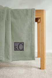 Grüne Kuscheldecke von s.Oliver, drapiert über eine Holzkante, mit sichtbarem Logo. Perspektive von der Seite.