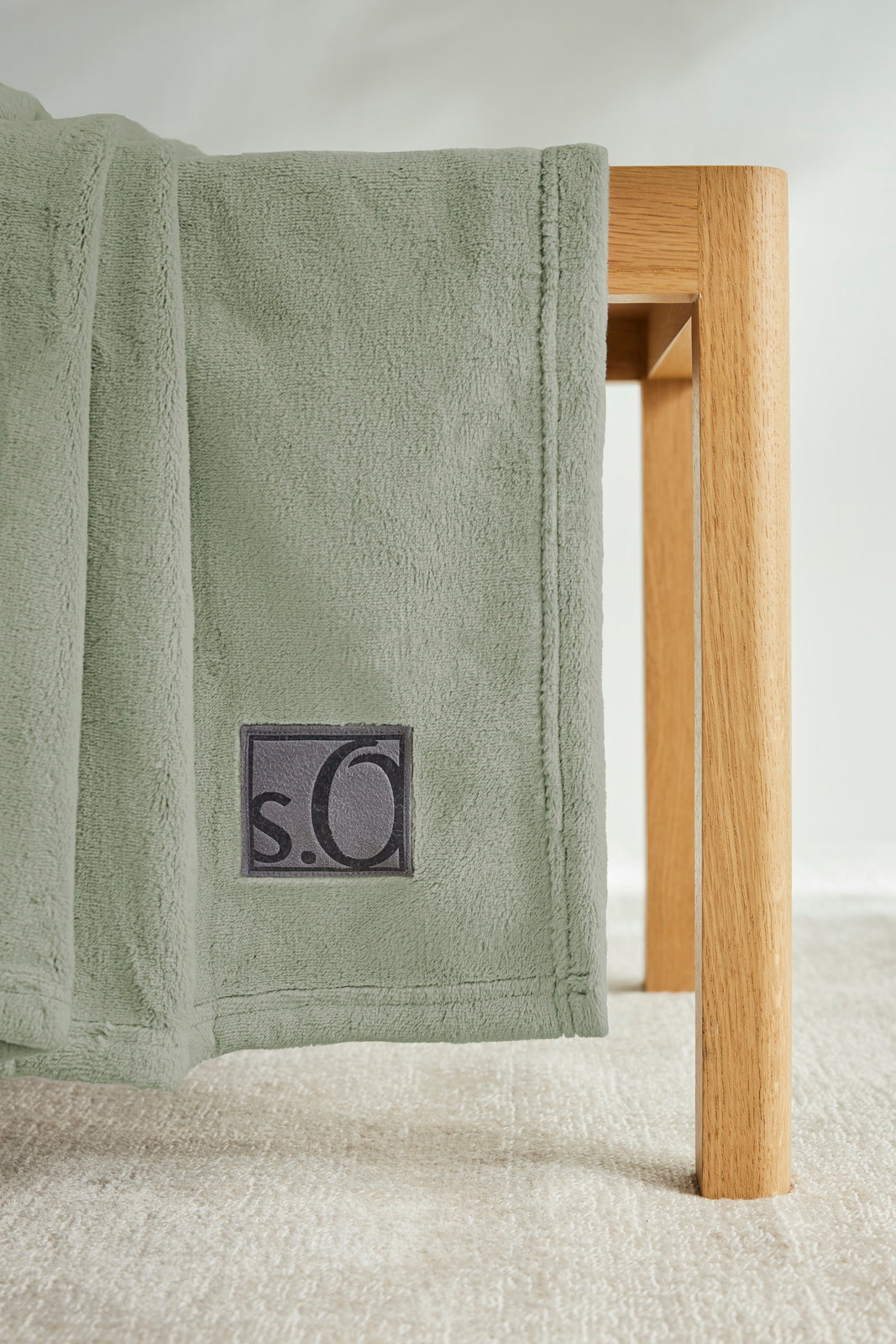 Grüne Kuscheldecke von s.Oliver, drapiert über eine Holzkante, mit sichtbarem Logo. Perspektive von der Seite.