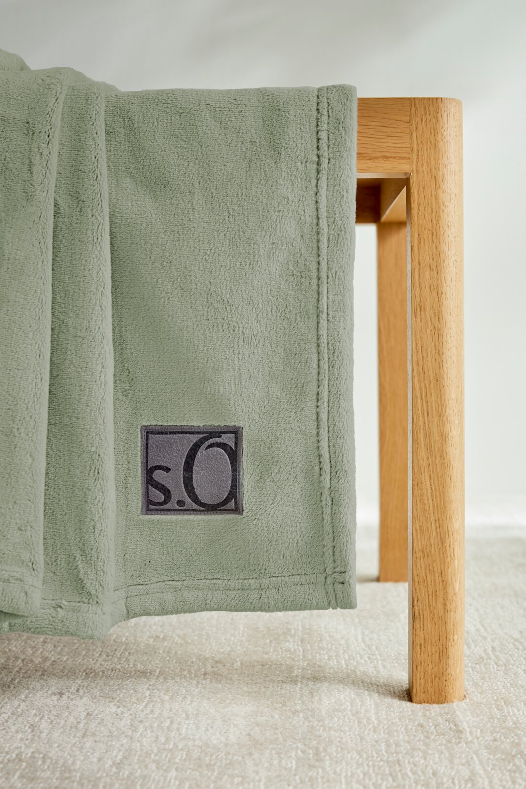 Grüne Kuscheldecke von s.Oliver, drapiert über eine Holzkante, mit sichtbarem Logo. Perspektive von der Seite.