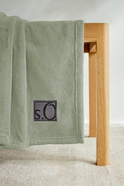 Grüne Kuscheldecke von s.Oliver, drapiert über eine Holzkante, mit sichtbarem Logo. Perspektive von der Seite.