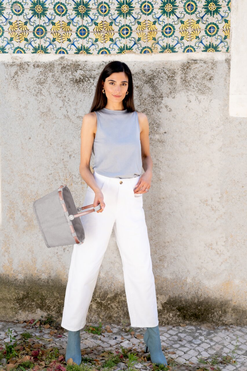 Frau in weißer Hose und grauem Top hält einen grauen carrybag XS in der Hand, seitliche Perspektive.