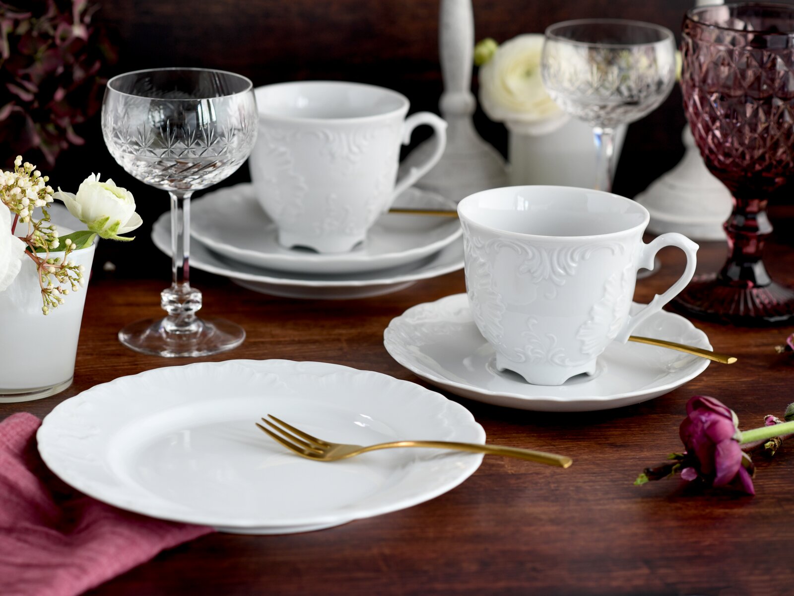 Elegantes weißes Porzellan-Kaffeegedeck mit reliefiertem Dekor (Tasse auf Untertasse) und passendem Teller auf Holztisch, mit goldfarbenem Besteck und Kristallgläsern im Hintergrund, fotografiert aus leicht erhöhter Frontperspektive
