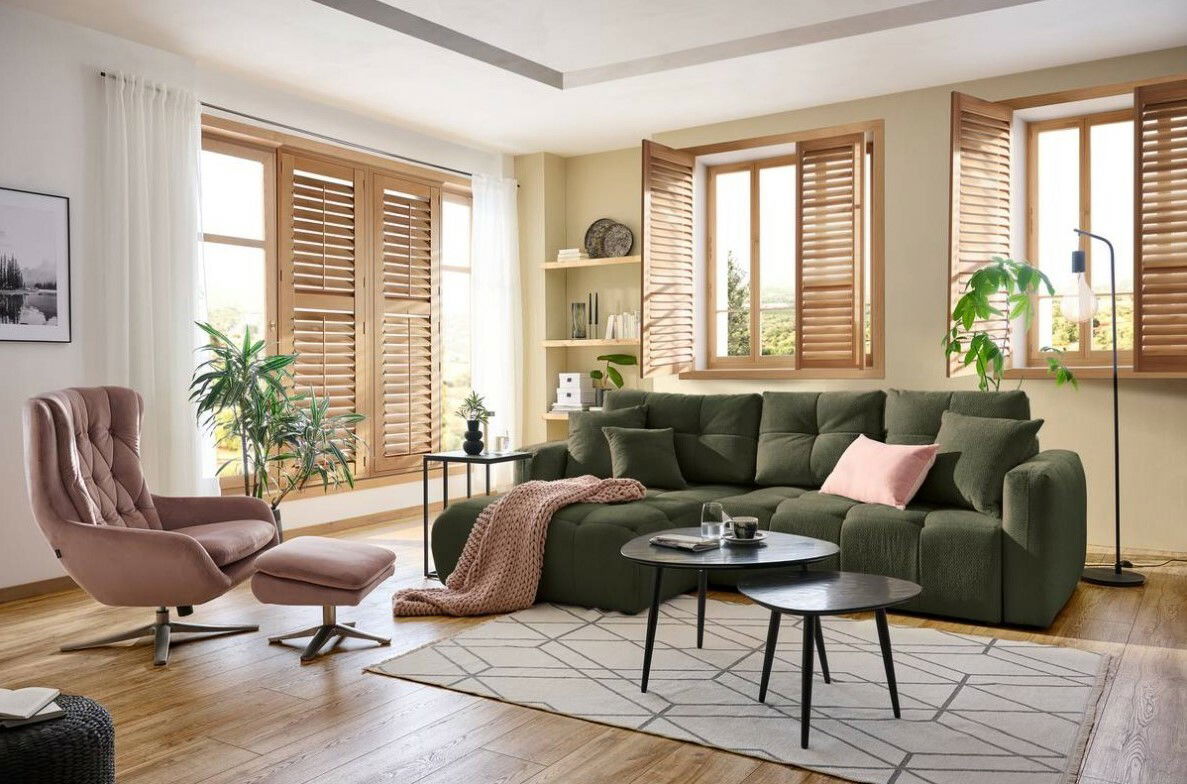 Gemütliches Wohnzimmer mit einer großen, grünen Rundecke im Vordergrund, umgeben von Holzmöbeln und Pflanzen. Die Perspektive zeigt den Raum von vorne, mit einem Sessel und Hocker links im Bild.