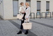 Een vrouw en een kind staan buiten voor een modern gebouw. Beiden houden een beige boodschappenmand met handvatten vast. De vrouw draagt een lange jas en zwarte laarzen, terwijl het kind een vest en zwarte laarzen draagt. De manden worden vanuit een zijaa