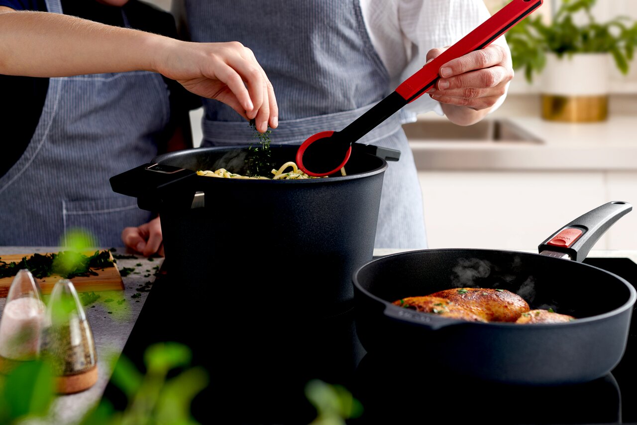 Zwei Personen kochen in einer modernen Küche. Im Vordergrund eine Guss-Hochrandpfanne mit gebratenem Fleisch, im Hintergrund ein Topf mit Nudeln. Perspektive von schräg oben.