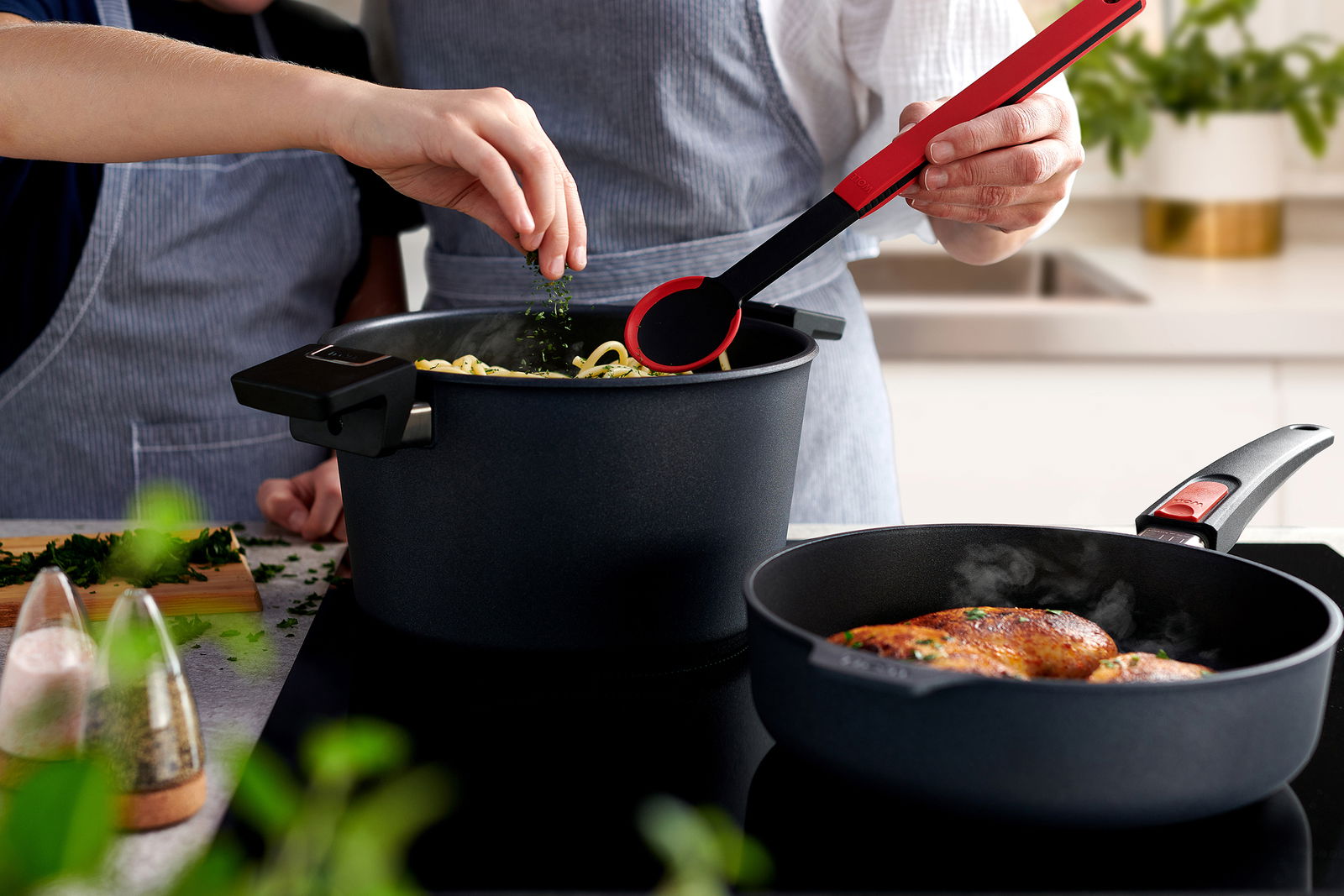Zwei Personen kochen in einer modernen Küche. Im Vordergrund eine Guss-Hochrandpfanne mit gebratenem Fleisch, im Hintergrund ein Topf mit Nudeln. Perspektive von schräg oben.