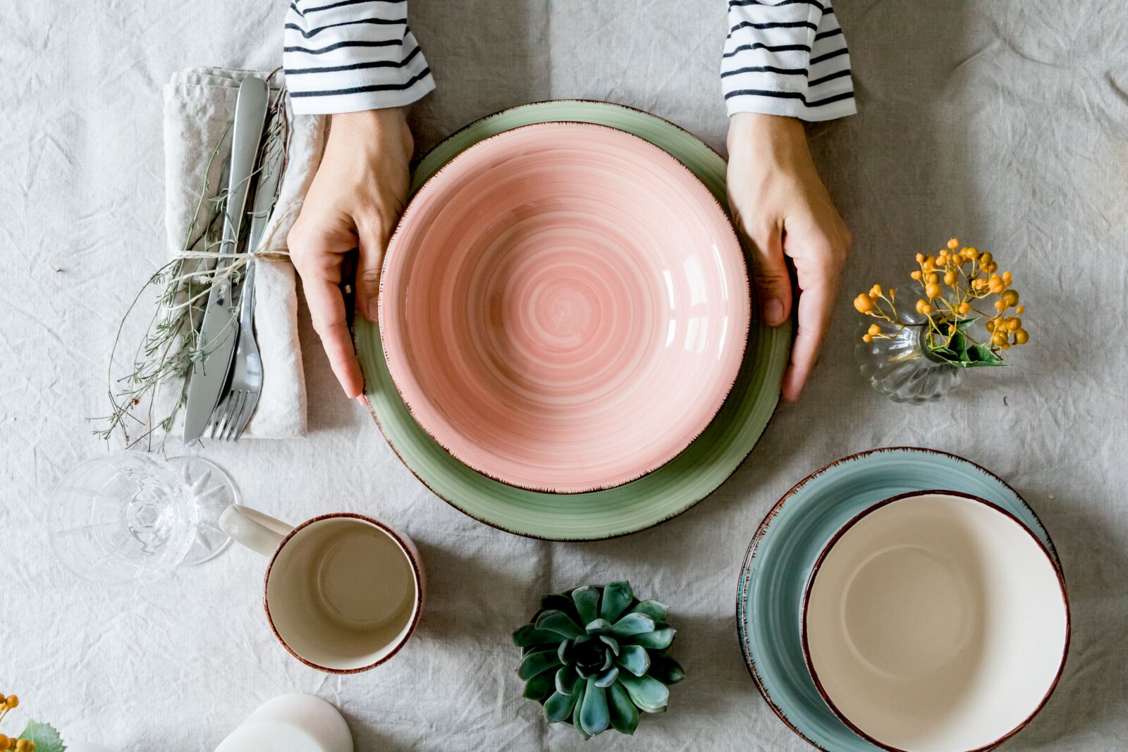 Draufsicht: Hände platzieren einen großen Keramikteller mit rosafarbenem Innenbereich und grünem Rand auf einer gedeckten Tischdecke, daneben Besteck, Tasse, Gläser und Schalen.