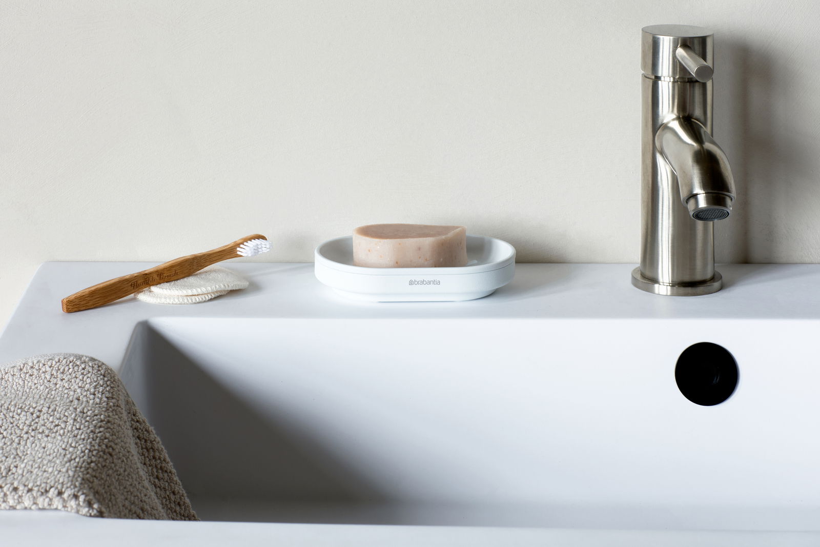 Seifenschale auf einem Waschbeckenrand mit Seife, Zahnbürste und Wasserhahn, aus der Perspektive von vorne betrachtet.