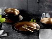 Schwarzes Geschirrset mit goldfarbenem Innenfinish (Schalen und Teller) auf dunklem Tisch, mit goldfarbenem Besteck und Gläsern, fotografiert in schräger Frontansicht von leicht oben.