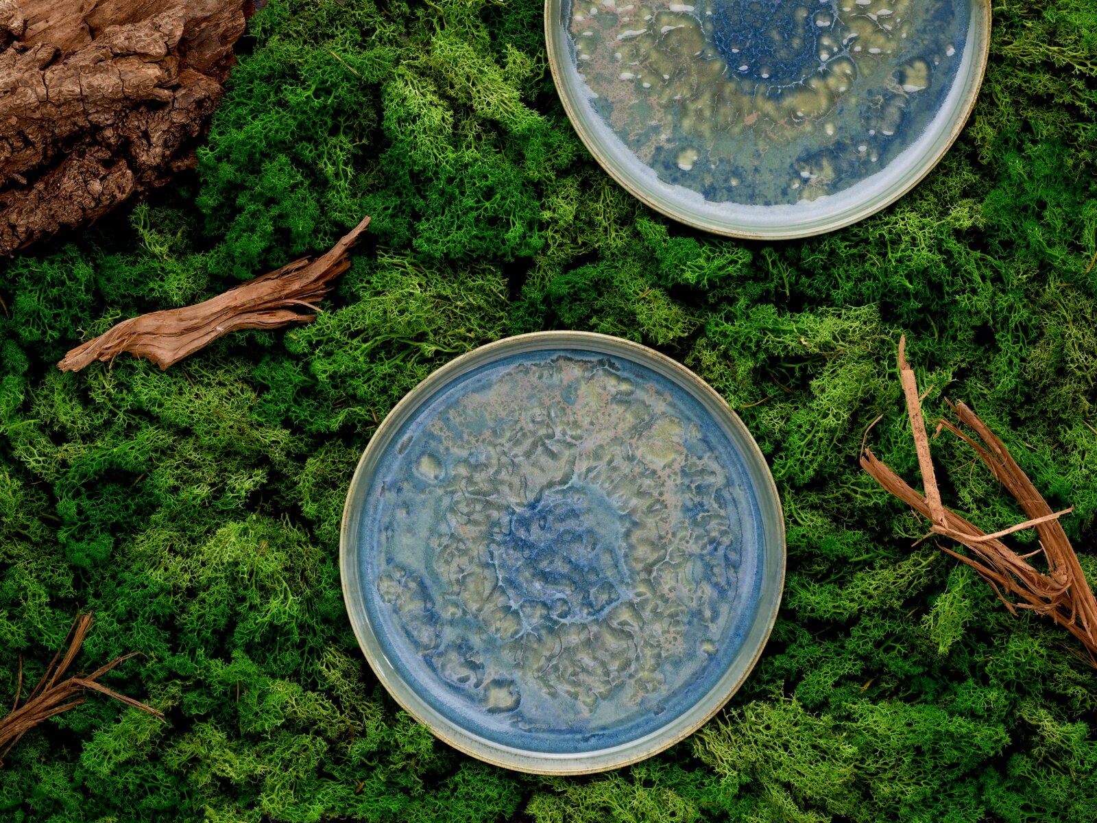 Draufsicht auf zwei runde Keramikteller mit blau-grün gesprenkelter Glasur auf moosigem Untergrund mit Holzstücken.
