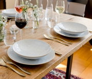 Schlichtes weißes Geschirr-Set mit tiefen Tellern auf rustikalem Holztisch, gedeckt mit Besteck, Weingläsern und kleinen Blumenvasen; Perspektive: leicht von oben in schräger Seitenansicht