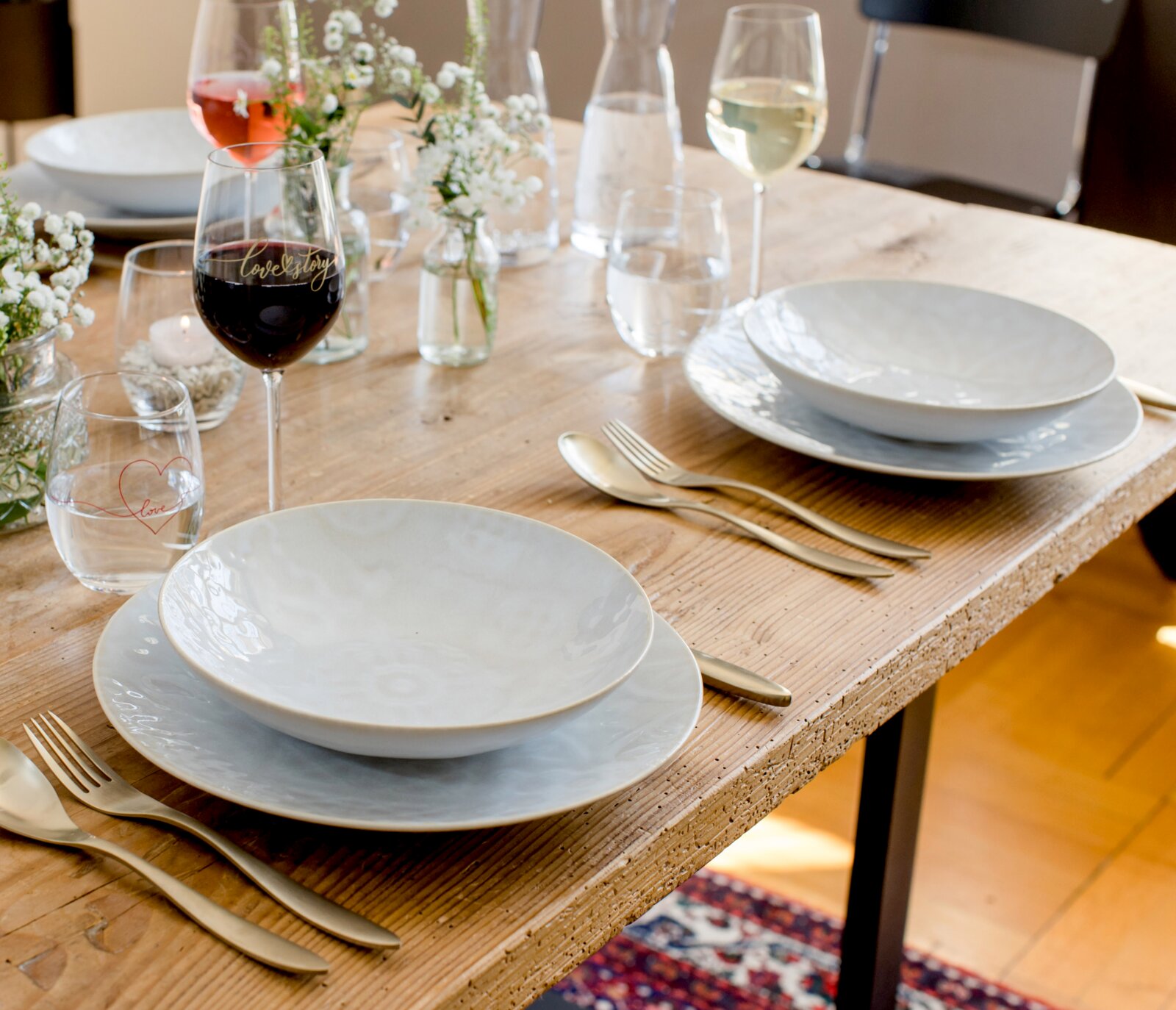 Schlichtes weißes Geschirr-Set mit tiefen Tellern auf rustikalem Holztisch, gedeckt mit Besteck, Weingläsern und kleinen Blumenvasen; Perspektive: leicht von oben in schräger Seitenansicht
