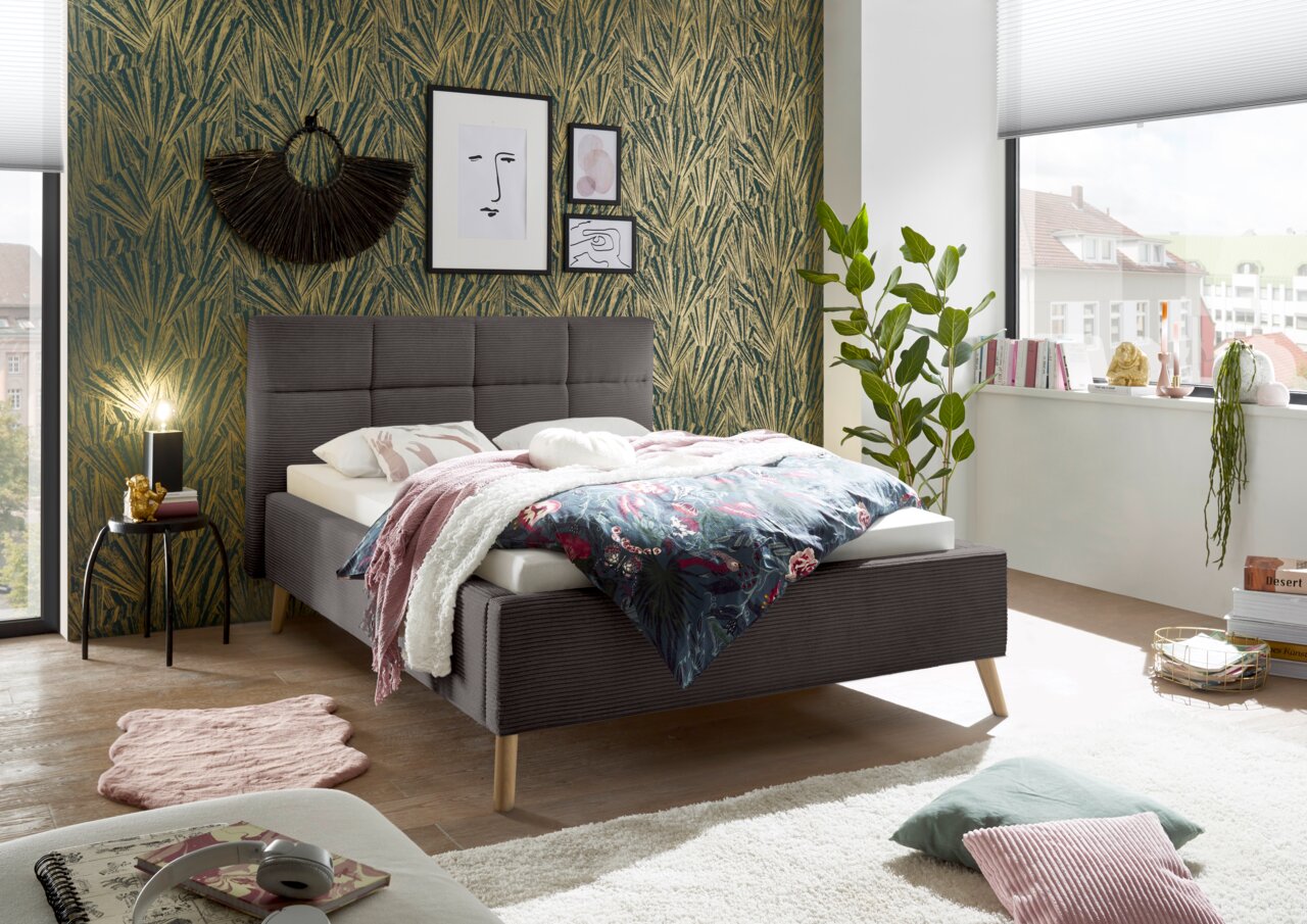 Modern gestoffeerd bed in een stijlvol ingerichte slaapkamer, zijaanzicht, met sierkussens en dekens.