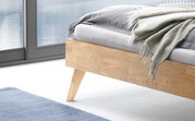 Nahaufnahme eines modernen Bettgestells aus hellem Holz mit schrägen Beinen, von der Seite fotografiert. Auf dem Bett liegt eine graue Decke.