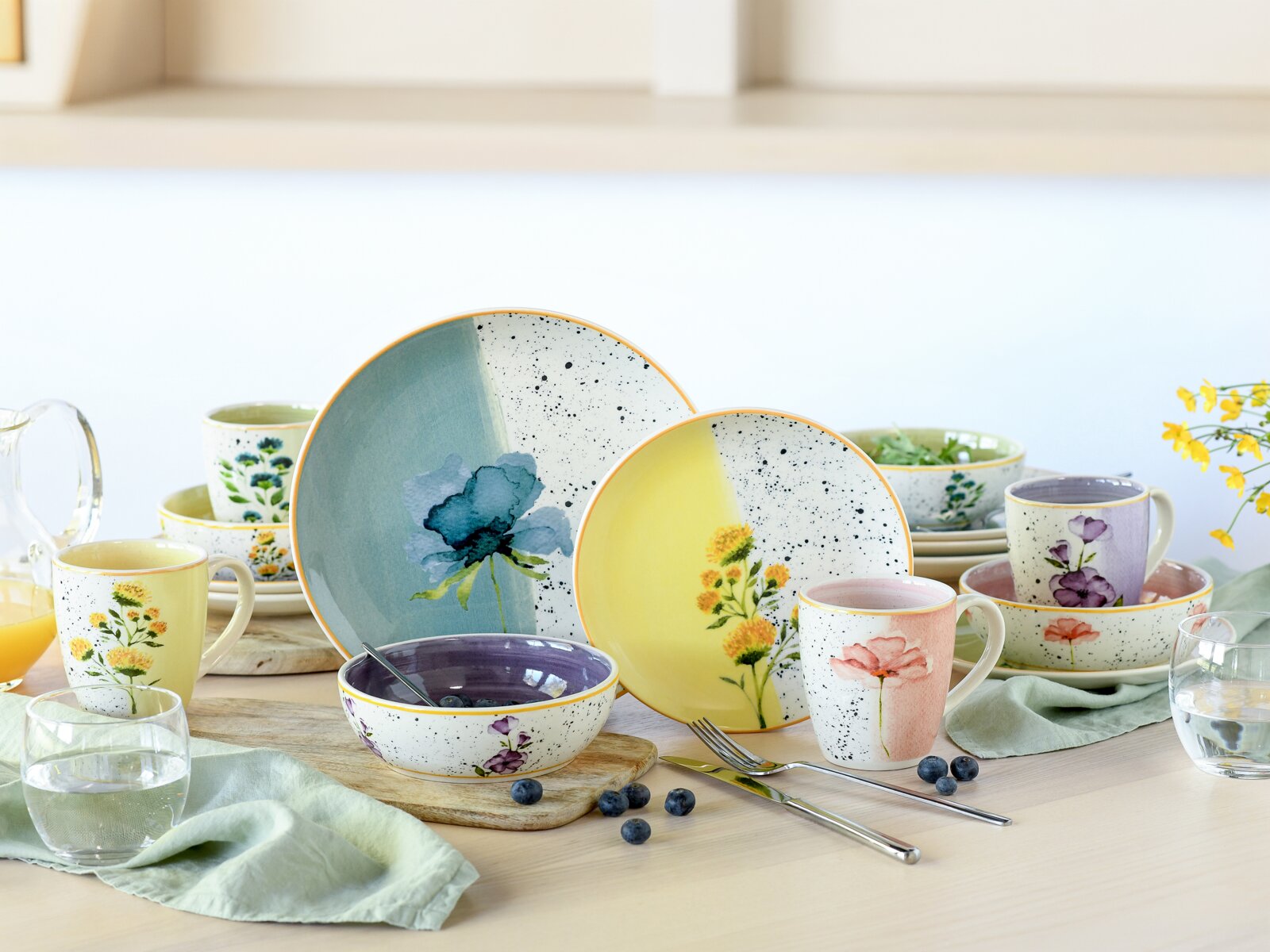 Farbenfrohes Keramikgeschirr mit floralen Aquarellmotiven, gesprenkeltem Dekor und orangefarbenen Rändern, arrangiert mit Tellern, Schalen und Tassen in frontaler Perspektive.