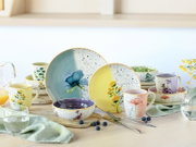 Farbenfrohes Keramikgeschirr mit floralen Aquarellmotiven, gesprenkeltem Dekor und orangefarbenen Rändern, arrangiert mit Tellern, Schalen und Tassen in frontaler Perspektive.