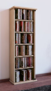 Schmales, hohes CD-Regal aus hellem Holz mit mehreren Einlegeböden, gefüllt mit CDs, freistehend auf Holzboden vor weißer Wand; Ansicht schräg von vorn links.