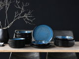 Elegantes 18-teiliges Tellerset in Blau, präsentiert auf einem Tisch aus der Seitenperspektive, mit einem modernen, dunklen Design und einem dekorativen Ast im Hintergrund.