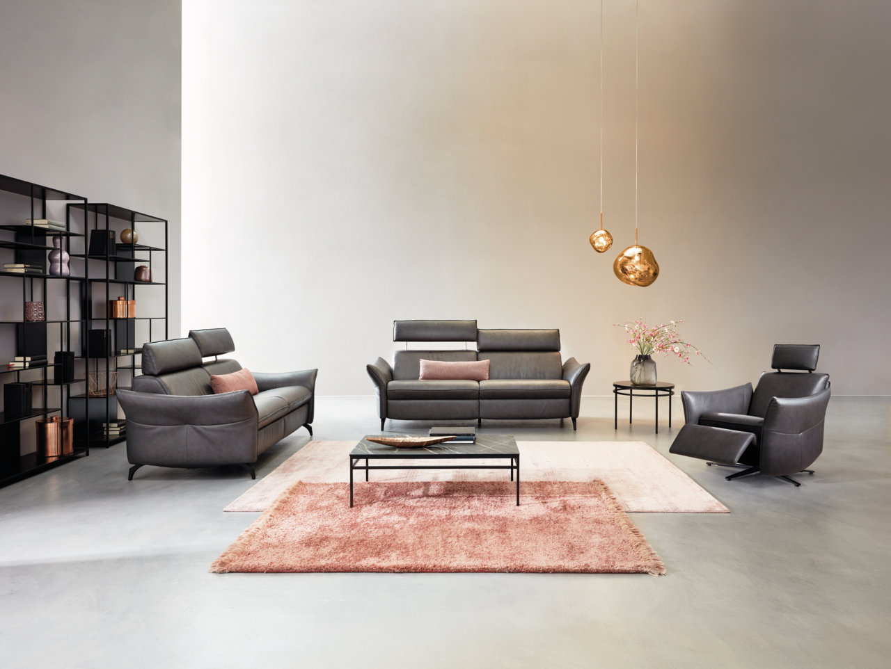 Wohnzimmeransicht mit Himolla Sofa PLANOPOLY MOTION, bestehend aus einem 3er-Sofa, einem 2,5er-Sofa und einem Sessel in dunklem Leder. Moderne Einrichtung mit Teppichen, Couchtisch und dekorativen Hängelampen.