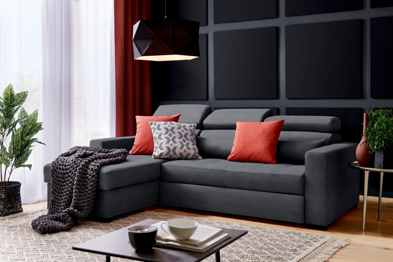 Graues Ecksofa mit roten und gemusterten Kissen, dekoriert mit einer Decke, in einem modernen Wohnzimmer mit Pflanzen und einem Couchtisch, seitliche Perspektive.
