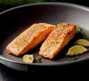 Twee goudbruin gebakken zalmfilets met kruidenkorst in een zwarte koekenpan, met partjes limoen en dille ernaast, gefotografeerd in schuin bovenaanzicht.