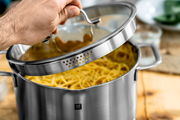 Nahaufnahme eines Edelstahlkochtopfs mit Glasdeckel, der von einer Hand geöffnet wird. Der Topf ist mit Spaghetti gefüllt und hat ein integriertes Abtropfsieb im Deckelrand.