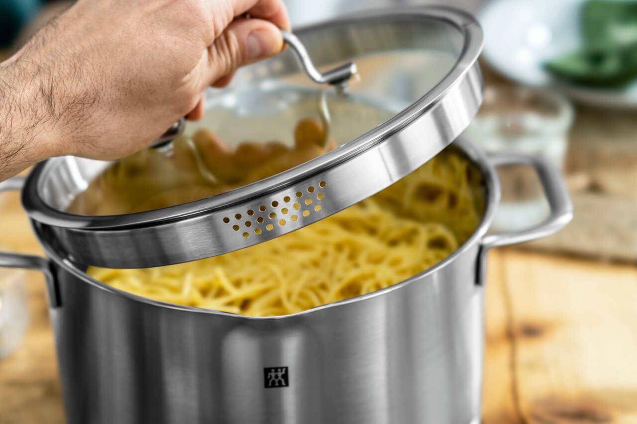 Close-up van een roestvrijstalen pan met glazen deksel, die met één hand wordt geopend. De pan is gevuld met spaghetti en heeft een ingebouwd vergiet in de rand van het deksel.