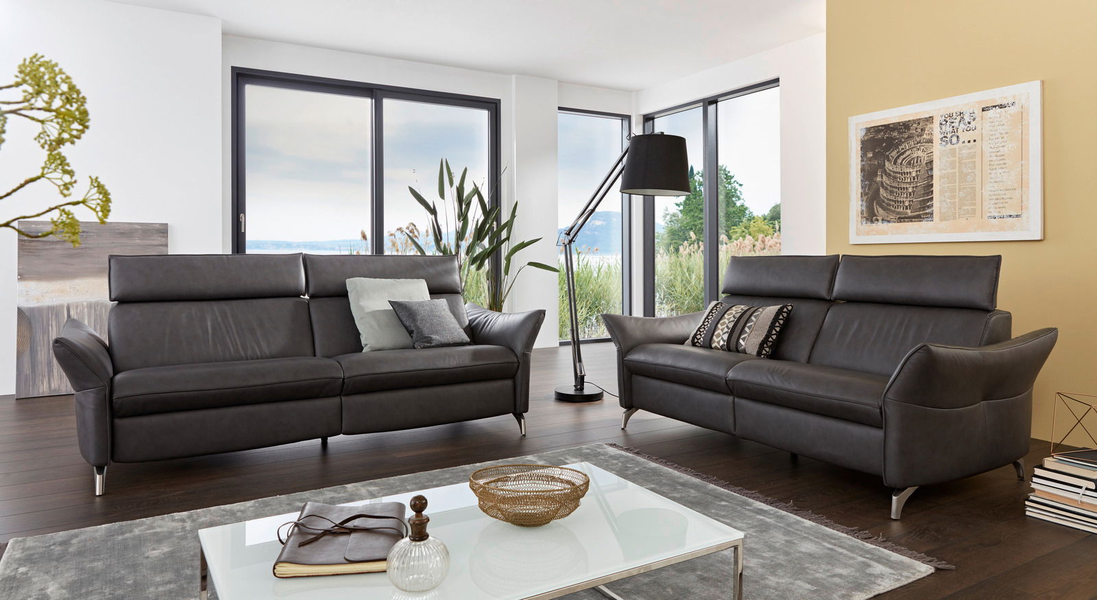 Zwei moderne, dunkelgraue Ledersofas in einem hellen Wohnzimmer mit großen Fenstern und Blick ins Grüne. Perspektive von vorne.